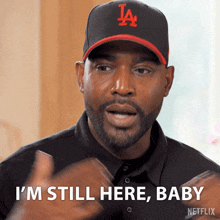 A man wearing a black LA cap and black shirt gestures confidently while saying "I'M STILL HERE, BABY" in bold text. The expression and gesture emphasize determination and resilience, creating a bold and self-assured vibe. The GIF is from Netflix.
