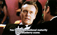 A man in a tuxedo delivers a cutting remark, saying, "You have the emotional maturity of a blueberry scone." His expression is calm yet disdainful, emphasizing the insult's humor and sophistication. The setting appears formal, with the line adding a touch of witty sarcasm to the interaction.