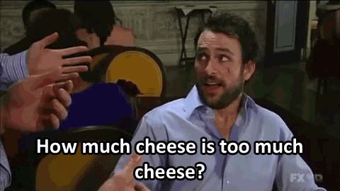 A man with a puzzled expression is sitting at a table, asking, "How much cheese is too much cheese?" while looking at someone off-screen. His tone suggests he's serious but also a bit confused or overwhelmed by the question.