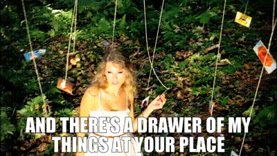 A woman in a white dress stands in a forested setting with photos hanging around her on strings. The text on the GIF reads, "And there's a drawer of my things at your place," referencing lyrics from a song. Her expression is soft and nostalgic, fitting the romantic, sentimental atmosphere created by the surrounding nature and suspended photographs.