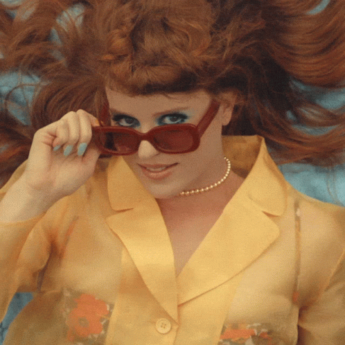 
A woman with red hair lies down while pulling her red sunglasses down to give a playful, knowing look. She is wearing a yellow blouse and her expression is confident and flirty.