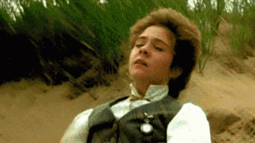 The image shows a scene from the movie A Room with a View featuring a young man, George Emerson (played by Julian Sands), lying on the ground in a meadow. He appears to be in a moment of emotional intensity or contemplation. His clothes are formal, matching the early 20th-century setting of the film. The greenery of the surroundings contrasts with his still and reflective pose, emphasizing a pivotal moment in the story.