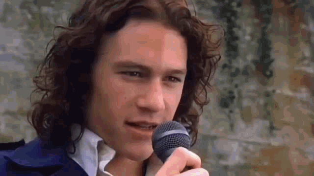 The image is from the movie 10 Things I Hate About You featuring Heath Ledger as Patrick Verona. He is holding a microphone, smiling slightly, and looking confident. This is from the famous scene where Patrick serenades Kat Stratford on the school bleachers, singing "Can't Take My Eyes Off You" in an effort to win her over. His charming and playful expression adds to the romantic atmosphere of the moment.