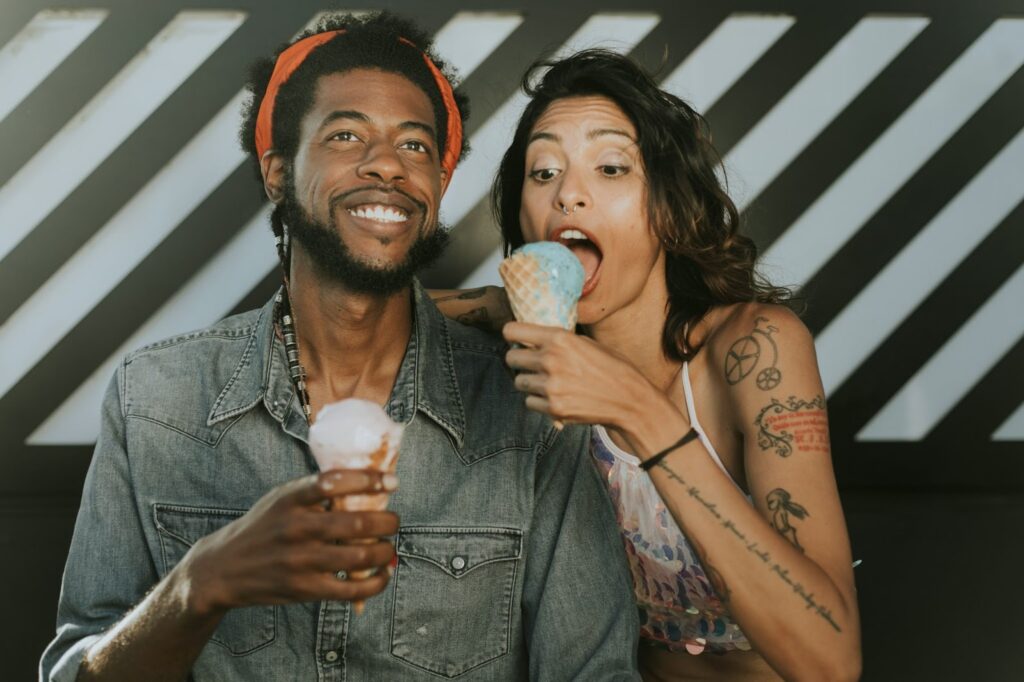 The image shows a cheerful couple enjoying ice cream cones together. The man, wearing a denim shirt and an orange headband, is smiling while holding his ice cream. The woman next to him, with tattoos and a colorful top, is enthusiastically eating her blue ice cream cone. They are sitting side by side in a casual, fun setting, capturing a playful and happy moment.