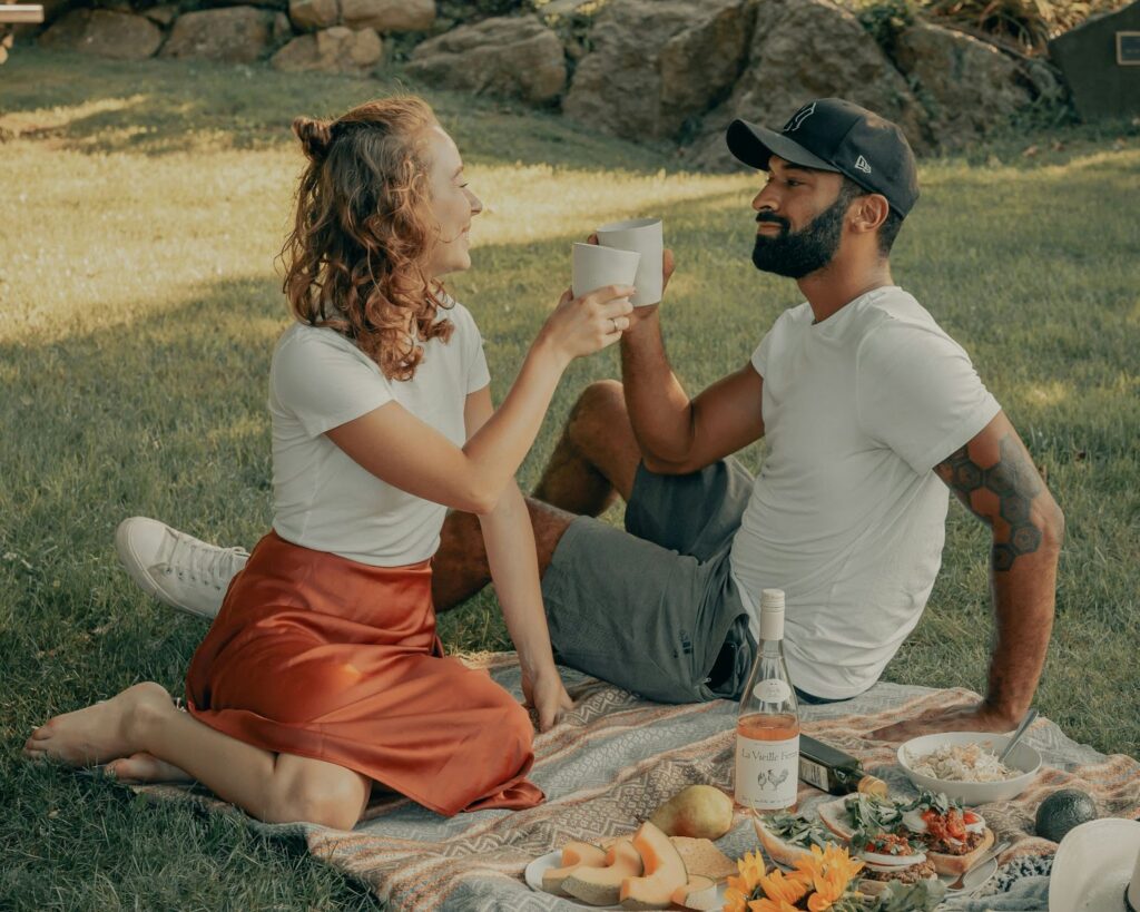 The image shows a couple enjoying a picnic outdoors, sitting on a blanket in a grassy area. They are smiling and clinking cups in a toast. The woman is wearing a white t-shirt and a rust-colored skirt, while the man is dressed casually in a white t-shirt, gray shorts, and a black cap. In front of them, the picnic spread includes a bottle of wine, fruits, and various snacks, creating a relaxed and joyful atmosphere.