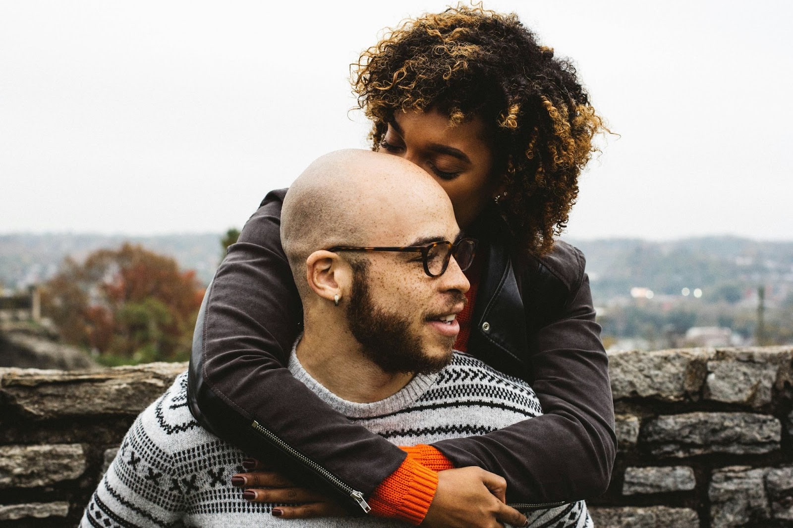 A couple shares an affectionate moment outdoors, with the woman embracing the man from behind and kissing the top of his head. The man, wearing glasses and a patterned sweater, looks off to the side with a gentle smile. The scene is warm and intimate, set against a soft, blurred background of nature, creating a cozy, loving atmosphere.