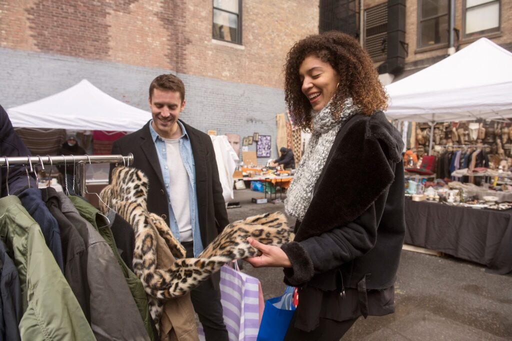 The image shows a couple shopping at an outdoor market, browsing through a rack of jackets. The woman is smiling as she holds up a leopard-print coat, while the man looks on with a smile. They are dressed warmly in coats and scarves, and various stalls and tents in the background display other items for sale. The atmosphere is casual and cheerful, suggesting they are enjoying a day of shopping together.