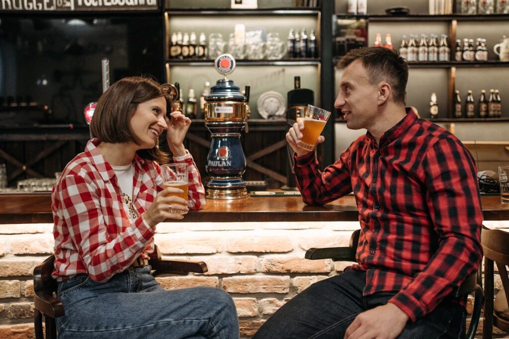 The image shows a couple sitting at a bar, enjoying drinks and laughing together. Both are dressed in casual, red plaid shirts. The woman, smiling and holding a glass of beer, brushes her hair back while the man raises his own beer glass as they engage in conversation. Behind them is a bar setup with taps and shelves stocked with bottles, creating a relaxed and cozy atmosphere.