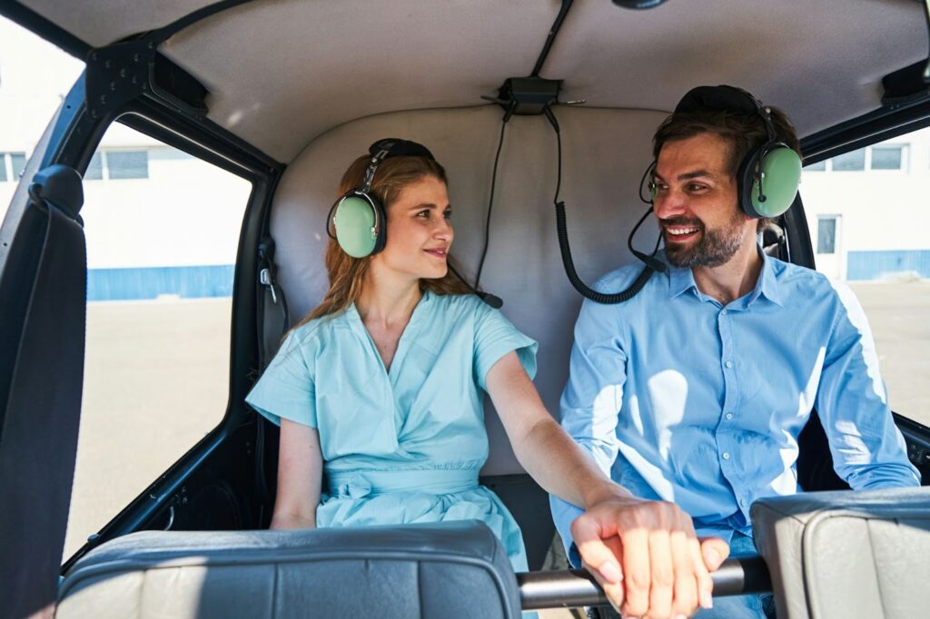 The image shows a couple sitting inside a helicopter, wearing headsets with microphones. They are smiling at each other and holding hands, creating a warm and affectionate atmosphere. The man is dressed in a light blue shirt, and the woman is wearing a light teal outfit, possibly a uniform. The scene suggests they are about to embark on a flight or have just finished one, sharing a happy moment together in the cockpit.