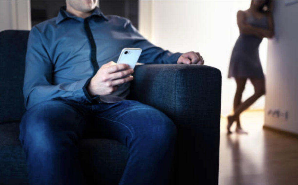 A man sits on a couch, focused on his phone, while a woman stands in the background, leaning against a wall with a distant expression. The image conveys a sense of emotional distance or tension between the two individuals.