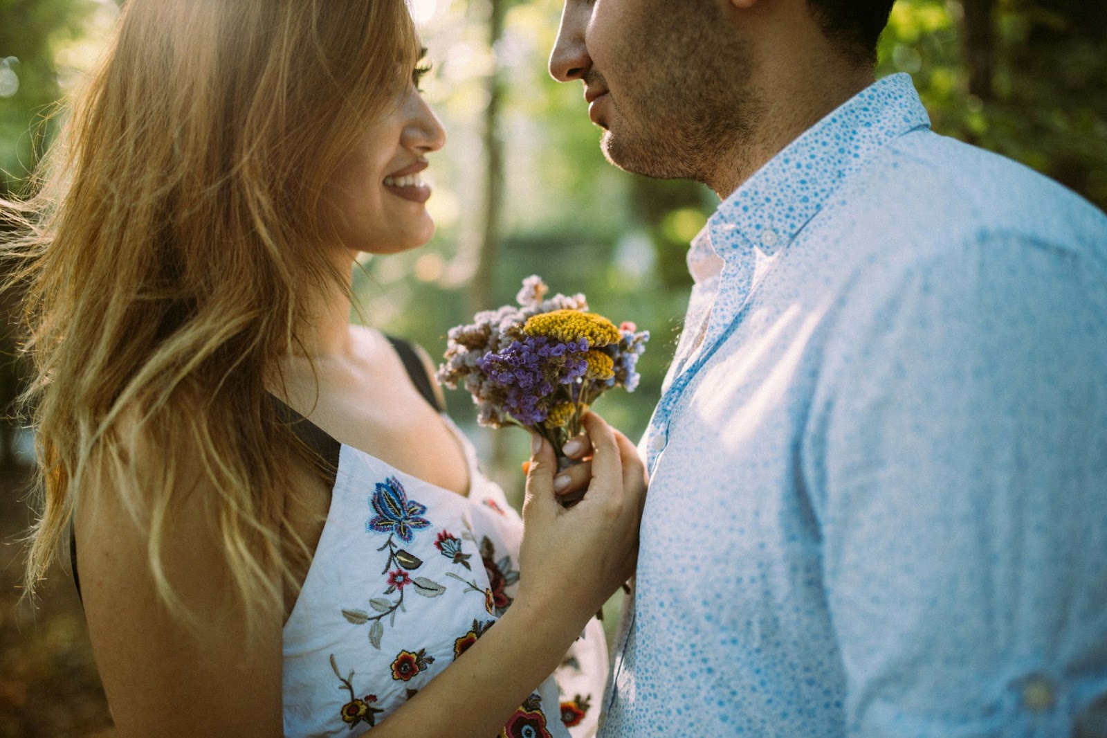 The image shows a young couple standing closely, facing each other in a romantic moment. The woman is smiling, holding a small bouquet of wildflowers near the man's chest. Both are casually dressed, and the setting appears to be outdoors in a sunlit, natural environment, possibly a forest or park.