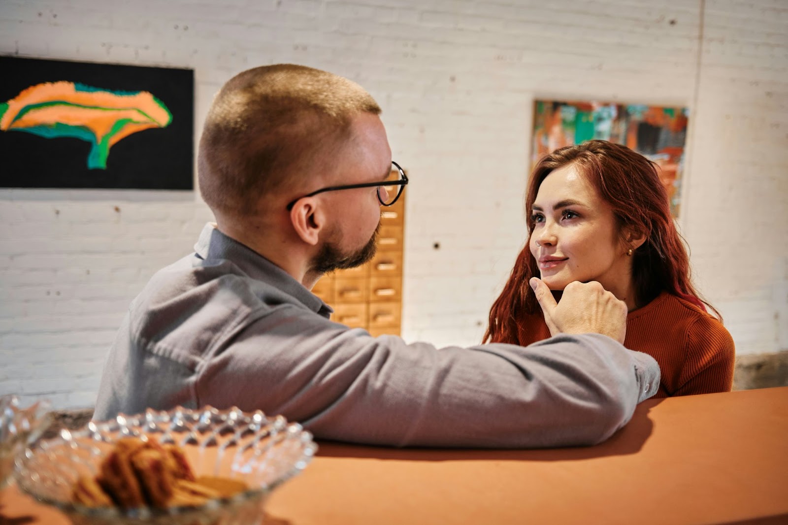 A man gently holds a woman's chin as they engage in a deep, intimate conversation. The woman gazes into his eyes with a soft expression, while they sit in a cozy, artistic setting with abstract art on the walls behind them.